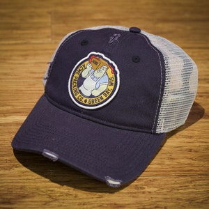 May include: A navy blue and white trucker hat with a distressed look. The hat features a circular patch with a cartoon character carrying a box and the text "Acme Packing Co. Green Bay, Wis."