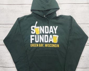 Könnte beinhalten: Dunkelgrüner Kapuzenpullover mit dem weißen Schriftzug "SUNDAY FUNDA" und zwei gelben Biergläsern. Darunter steht "GREEN BAY, WISCONSIN". Der Pullover hat eine Kapuze und eine Vordertasche.