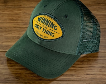 Könnte beinhalten: Grüne Trucker-Kappe mit einem gestickten Aufnäher in Gelb und Grün, auf dem "Winning, It's the Only Thing, Green Bay, Wis." steht.