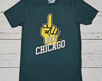 Könnte beinhalten: Ein dunkelgrünes Kurzarm-T-Shirt mit einem S-Größenetikett und dem Tundra Town-Logo am Halsausschnitt. Auf der Vorderseite befindet sich eine gelbe Schaumstofffinger-Grafik, die nach oben zeigt, mit dem Text HEY CHICAGO in weißen Buchstaben.