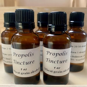 May include: Several amber glass bottles with black caps are arranged in a wooden tray. Each bottle has a white label with the words "Propolis Tincture" and "1 oz Wheat grain alcohol". The bottles are filled with a brown liquid.