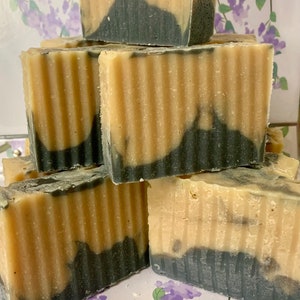 May include: Stacked bars of handmade soap with a two-tone design. The top portion is a light beige color, while the bottom half features a dark gray swirl. The soap bars have a textured, ridged surface. The background has a floral design.
