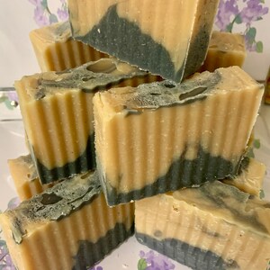 May include: A stack of handmade soap bars. Each bar has a light tan top and a dark gray bottom, with a wavy line separating the two colors. The bars are stacked on top of each other, creating a visually appealing display.