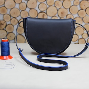 May include: A dark blue half-moon shaped leather crossbody bag with a blue and black strap. The bag is sitting on a light brown surface with a spool of blue thread.