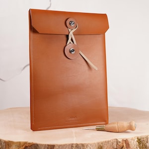 May include: A brown leather envelope-style laptop sleeve with a tan leather strap and metal button closure. The sleeve is sitting on a wooden surface with a leather awl next to it.