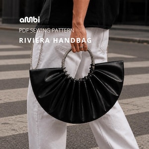 Op de afbeelding: Zwarte Riviera handtas met een geplooid ontwerp en een zilverkleurige handgreep. De tas heeft een kettingriem. De tekst "amBi" en "RIVIERA HANDBAG" zijn zichtbaar.