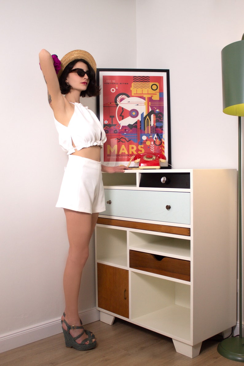 K&ouml;nnte beinhalten: Eine Person in einem wei&szlig;en Neckholder-Top und Shorts, mit Sonnenbrille und Strohhut. Ein farbenfrohes Poster mit dem Wort "Mars" h&auml;ngt &uuml;ber einem Schrank im Mid-Century-Modern-Stil mit Schubladen und offenen Regalen. Ein rotes W&auml;hlscheibentelefon steht oben.