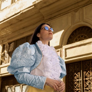 May include: A light blue jacket with puff sleeves and a textured floral pattern worn over a white shirt featuring a prominent ruffled front. The outfit is paired with a plain yellow skirt. The ensemble is completed by round sunglasses with dark lenses.