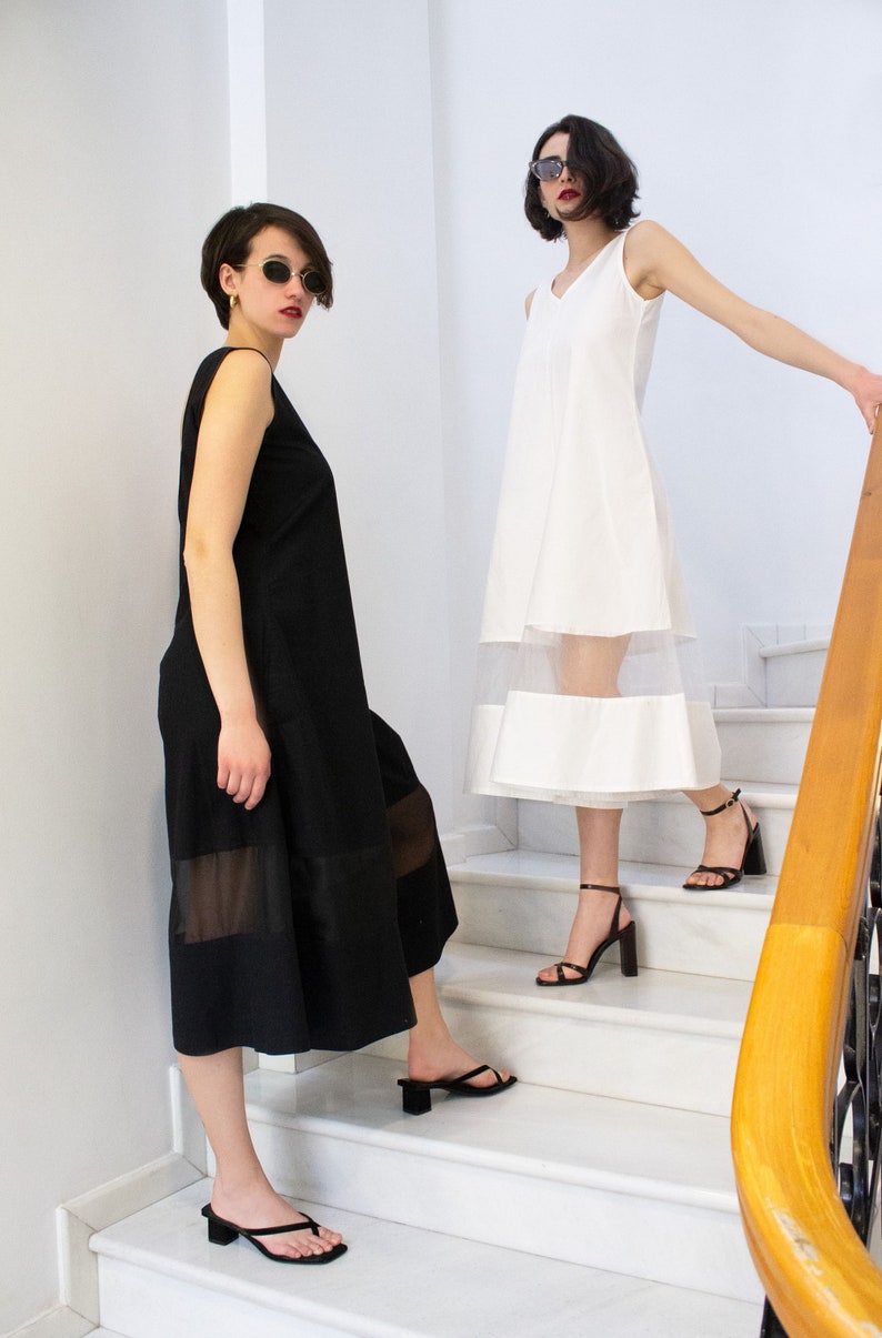 May include: Two women wearing black and white dresses with sheer panels stand on a white marble staircase. Both women are wearing sunglasses and black heeled sandals.
