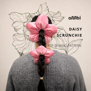May include: Two pink satin flower-shaped scrunchies worn in a braided hairstyle. The text reads: ambi DAISY SCRUNCHIE PDF SEWING PATTERN. The background features a black line drawing of a large flower, and the wearer is in a grey sweater.