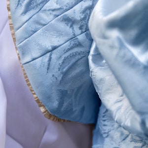 May include: A light blue fabric with a tonal floral jacquard pattern and a subtle sheen. The material features structured pleats and a beige pleated trim along the curved edge, layered over a smooth, solid white fabric base.