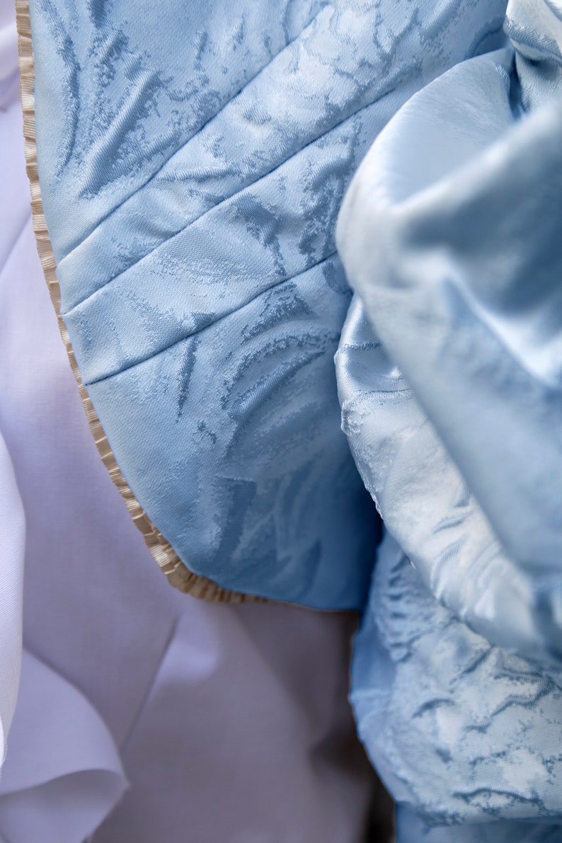 May include: A light blue fabric with a tonal floral jacquard pattern and a subtle sheen. The material features structured pleats and a beige pleated trim along the curved edge, layered over a smooth, solid white fabric base.