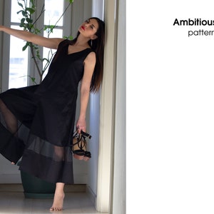 May include: A woman wearing a black sleeveless dress with sheer panels and a V-neckline. She is standing in a hallway and holding a pair of black heeled sandals. The text "Ambitious elle patterns" is visible in the upper right corner of the image.