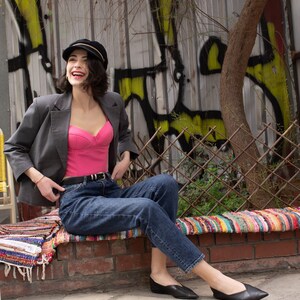 Peut inclure: Une femme portant un blazer gris, un haut corset rose, un jean bleu et des chaussures noires est assise sur un mur en briques avec un tapis color&eacute; devant elle. Elle sourit et regarde sur le c&ocirc;t&eacute;.
