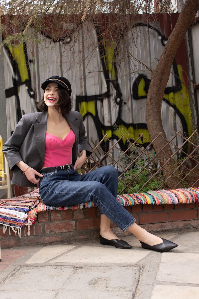 Peut inclure: Une femme portant un blazer gris, un haut corset rose, un jean bleu et des chaussures noires est assise sur un mur en briques avec un tapis color&eacute; devant elle. Elle sourit et regarde sur le c&ocirc;t&eacute;.