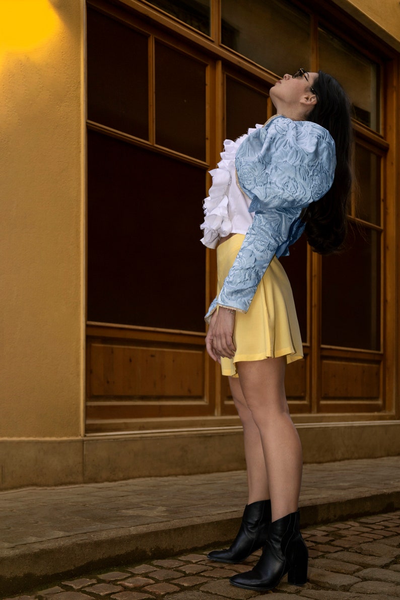 May include: A long-sleeved top featuring light blue textured fabric with puff sleeves, a white ruffled front panel, and a solid yellow mini skirt. The outfit is paired with black leather ankle boots with a block heel and a side zipper closure.