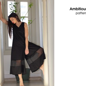 May include: A black jumpsuit with sheer panels at the bottom. The jumpsuit has a v-neck and sleeveless design. The model is standing in a hallway with a plant in the background.