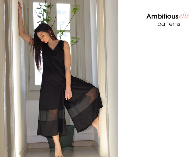 May include: A black jumpsuit with sheer panels at the bottom. The jumpsuit has a v-neck and sleeveless design. The model is standing in a hallway with a plant in the background.