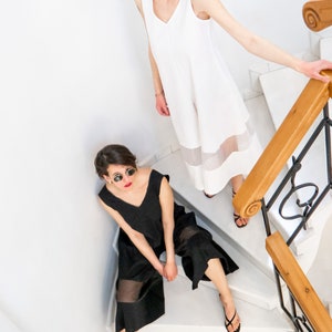 May include: Two women wearing sunglasses and sleeveless dresses with sheer panels stand on a white staircase. The woman on the top of the stairs is wearing a white dress and the woman on the bottom of the stairs is wearing a black dress.