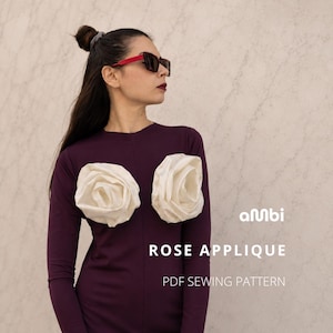 May include: A long-sleeved dark purple top featuring two large, white, three-dimensional fabric rose appliques on the chest. The text "ambi ROSE APPLIQUE PDF SEWING PATTERN" is printed in white on the right side of the image.
