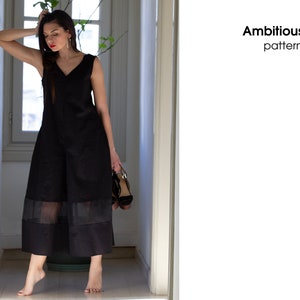 May include: A woman wearing a black sleeveless jumpsuit with a sheer overlay at the hem. The jumpsuit has a V-neckline and is made of a lightweight fabric. The woman is standing in a hallway with a window in the background. The text "Ambitious elle patterns" is visible in the image.