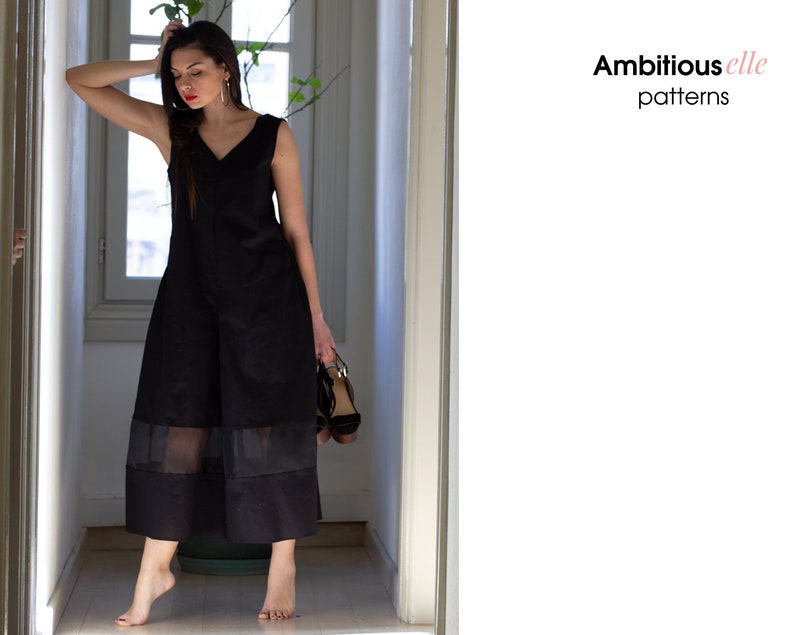 May include: A woman wearing a black sleeveless jumpsuit with a sheer overlay at the hem. The jumpsuit has a V-neckline and is made of a lightweight fabric. The woman is standing in a hallway with a window in the background. The text "Ambitious elle patterns" is visible in the image.