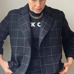 Peut inclure: Un blazer noir &agrave; motif &agrave; carreaux blancs. Le blazer a un col crant&eacute; et des manches longues. La personne porte un t-shirt noir avec le texte "K C" imprim&eacute; dessus. La personne porte &eacute;galement un collier en cha&icirc;ne dor&eacute;e.