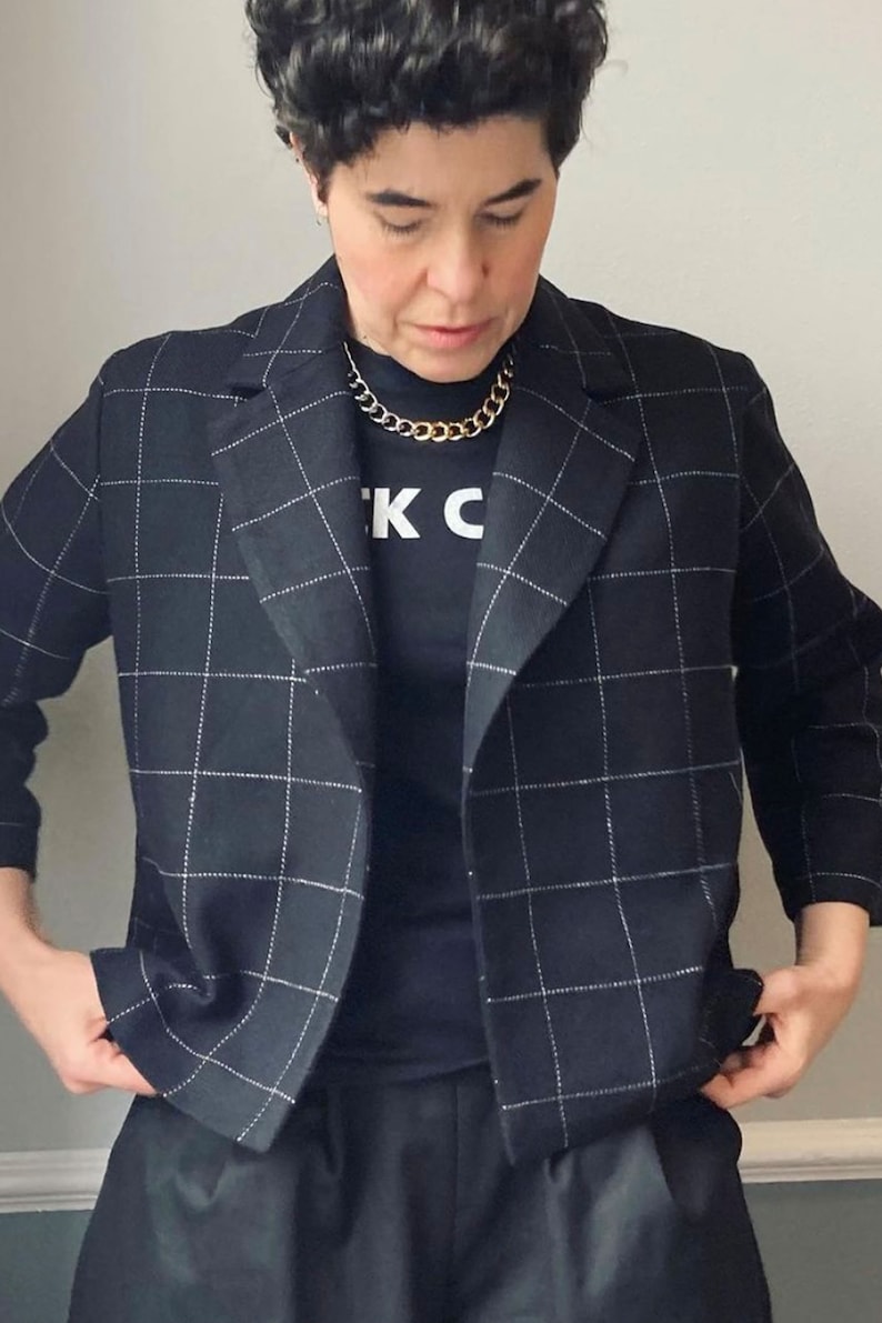 Peut inclure: Un blazer noir &agrave; motif &agrave; carreaux blancs. Le blazer a un col crant&eacute; et des manches longues. La personne porte un t-shirt noir avec le texte "K C" imprim&eacute; dessus. La personne porte &eacute;galement un collier en cha&icirc;ne dor&eacute;e.
