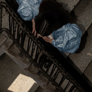 May include: A light blue blouse featuring voluminous puff sleeves with a textured, crinkled fabric finish. The garment has a fitted bodice and is worn by an individual with dark hair, viewed from a high-angle perspective above a staircase.