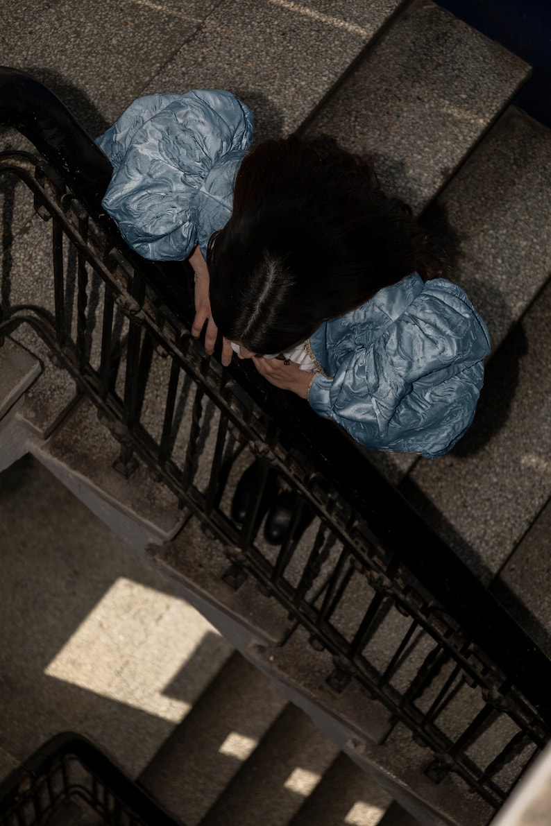May include: A light blue blouse featuring voluminous puff sleeves with a textured, crinkled fabric finish. The garment has a fitted bodice and is worn by an individual with dark hair, viewed from a high-angle perspective above a staircase.