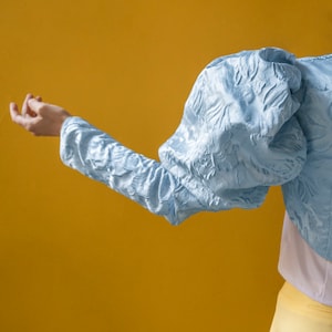 May include: A light blue cropped jacket featuring voluminous puff sleeves and a textured, raised floral pattern throughout the fabric. The garment is worn over a plain white top and a solid yellow skirt, showcasing a distinct silhouette.