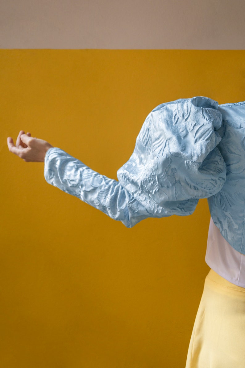 May include: A light blue cropped jacket featuring voluminous puff sleeves and a textured, raised floral pattern throughout the fabric. The garment is worn over a plain white top and a solid yellow skirt, showcasing a distinct silhouette.