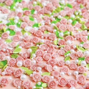 May include: A close-up of a pink and white background covered in small pink roses, green leaves, and white pearl beads.