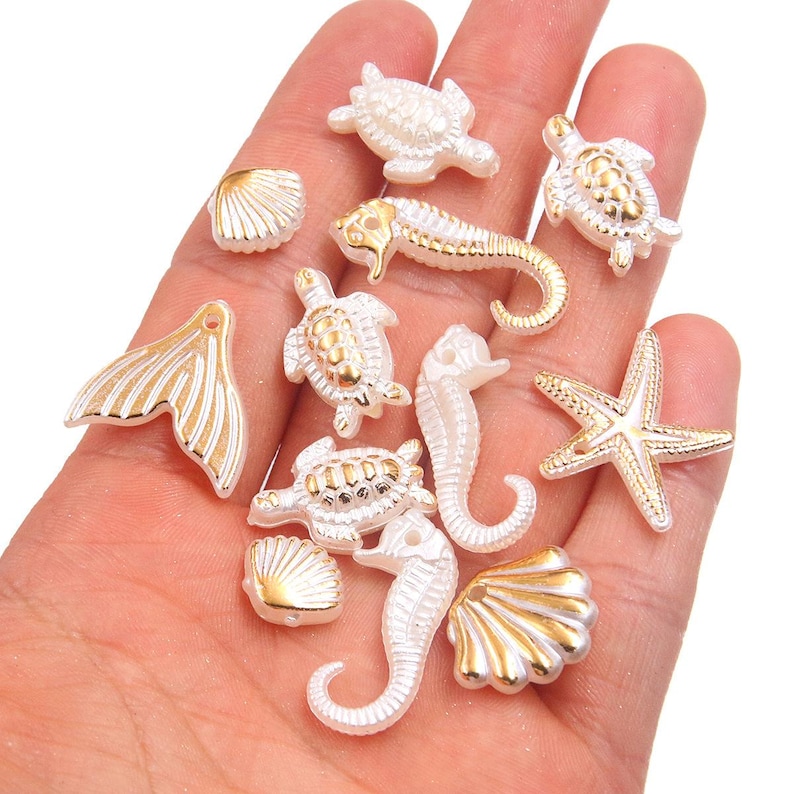 May include: A collection of white and gold-colored sea-themed charms, including starfish, seahorses, turtles, seashells, and a mermaid tail. These decorative charms are ideal for jewelry making or craft projects.