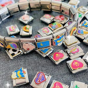 Puede incluir: Un brazalete de plata con dijes coloridos, que incluyen una cara de dibujos animados, un corazón con una huella de pata, una luna creciente con estrellas y un arcoíris. Los dijes están unidos a un brazalete de cadena de plata.