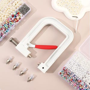 May include: A white hand press tool with a red handle, used for applying fasteners, sits next to several clear plastic containers filled with beads and pearls in various colors and sizes. A small tray of pearls and an Allen wrench are also visible.