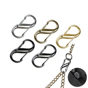 Puede incluir: Un surtido de cierres en forma de S en plata, oro y negro. Los cierres se muestran con una cadena dorada y una cremallera negra. Estos cierres se utilizan para la fabricación de joyas y otros proyectos de artesanía.