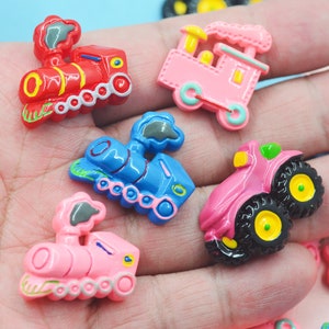 May include: Assortment of colorful plastic toy train and vehicle charms. The charms feature a red train, a blue train, a pink train, a pink truck, and a pink fire truck.