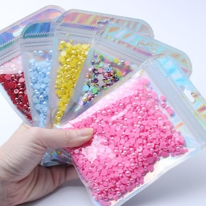 Può includere: Una mano che tiene diverse buste di plastica trasparente piene di perline colorate per l'artigianato. Le buste contengono perline rosse, blu, gialle, iridescenti e rosa. Le buste hanno una parte superiore olografica.