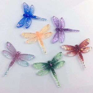 May include: Six colorful dragonfly ornaments in shades of blue, orange, purple, pink, green, and red. Each dragonfly has translucent wings with glitter accents and a detailed body. These decorative items are ideal for crafts or home decor.