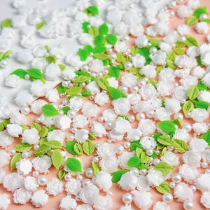 May include: A collection of white flower and leaf embellishments, perfect for crafting and decorating. The embellishments are scattered on a pink surface and include small white pearls.