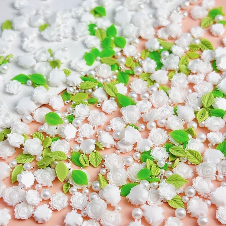 May include: A collection of white flower and leaf embellishments, perfect for crafting and decorating. The embellishments are scattered on a pink surface and include small white pearls.