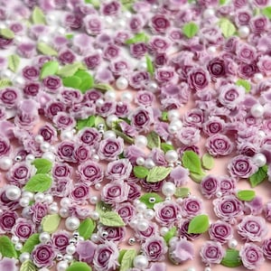 May include: A close-up of a large quantity of small, purple, and white flower embellishments. The flowers are made of polymer clay and are scattered on a pink background. There are also small, white pearl beads scattered throughout the image.