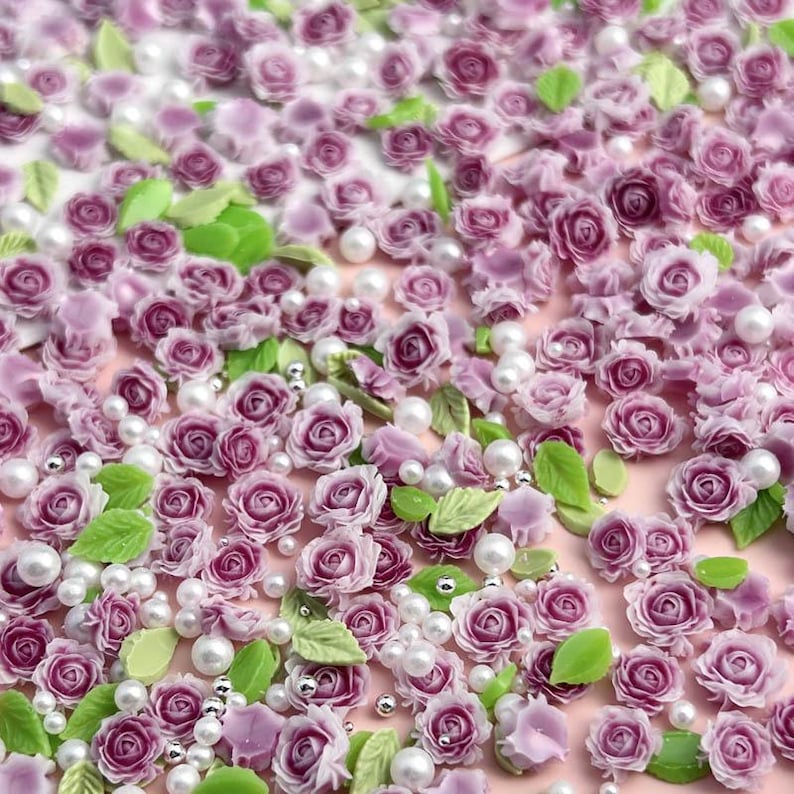 May include: A close-up of a large quantity of small, purple, and white flower embellishments. The flowers are made of polymer clay and are scattered on a pink background. There are also small, white pearl beads scattered throughout the image.