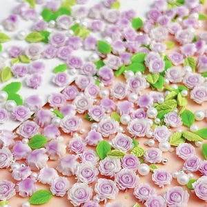 May include: A collection of small, purple and white 3D flower embellishments, perfect for nail art or crafting. The flowers are scattered on a white background with small white pearls.