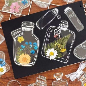May include: Clear, bottle-shaped stickers with white outlines and filled with dried flowers. One sticker has a sunflower, blue flowers, and the word "Believe". Another has a daisy and fern. Other stickers are also visible.