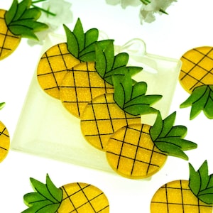 May include: A set of eight yellow and green pineapple shaped resin embellishments with glitter. The pineapples are arranged on a white surface.