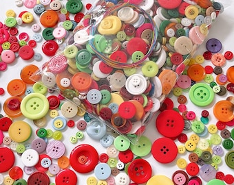 Puede incluir: Un gran surtido de botones coloridos de varios tamaños y formas, incluyendo rojo, verde, azul, amarillo y blanco. Los botones están en una bolsa de plástico transparente y esparcidos sobre una superficie blanca.