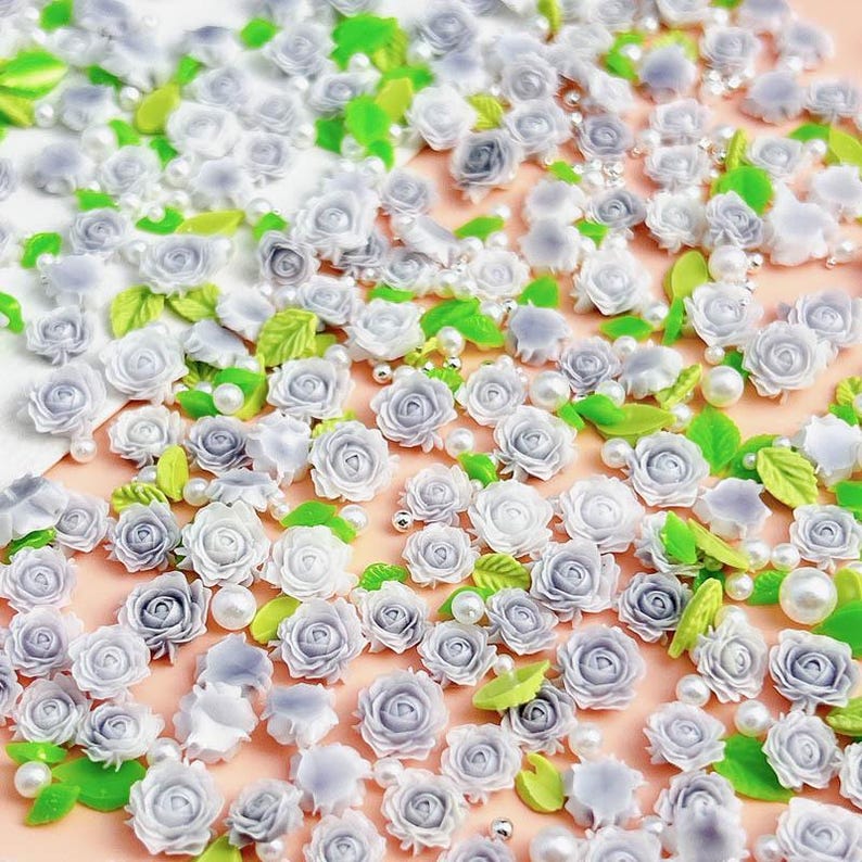 May include: A collection of small, white, and gray 3D polymer clay roses with green leaves and pearl beads. The roses are scattered on a pink surface.