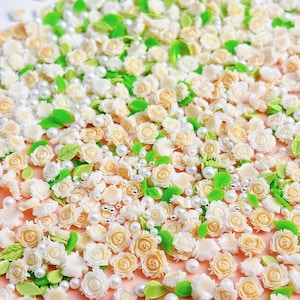 May include: A collection of small, white and cream-colored flower embellishments, perfect for crafting and decorating. The embellishments are scattered on a light pink surface, along with small white pearls.